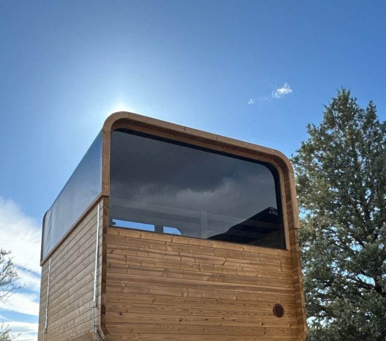 The rear window of a Thermory Sauna Square against a blue Oregon sky